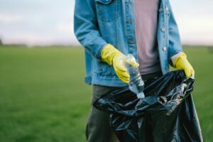 Un bénévole en veste en jean et gants ramasse une bouteille en plastique dans un sac poubelle dans un champ vert.