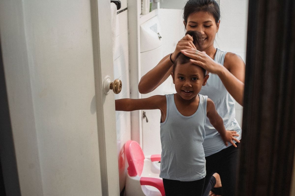 Une femme coiffe une petite fille souriante devant un petit pot rose dans une salle de bain Une femme coiffe une petite fille souriante devant un petit pot rose dans une salle de bain